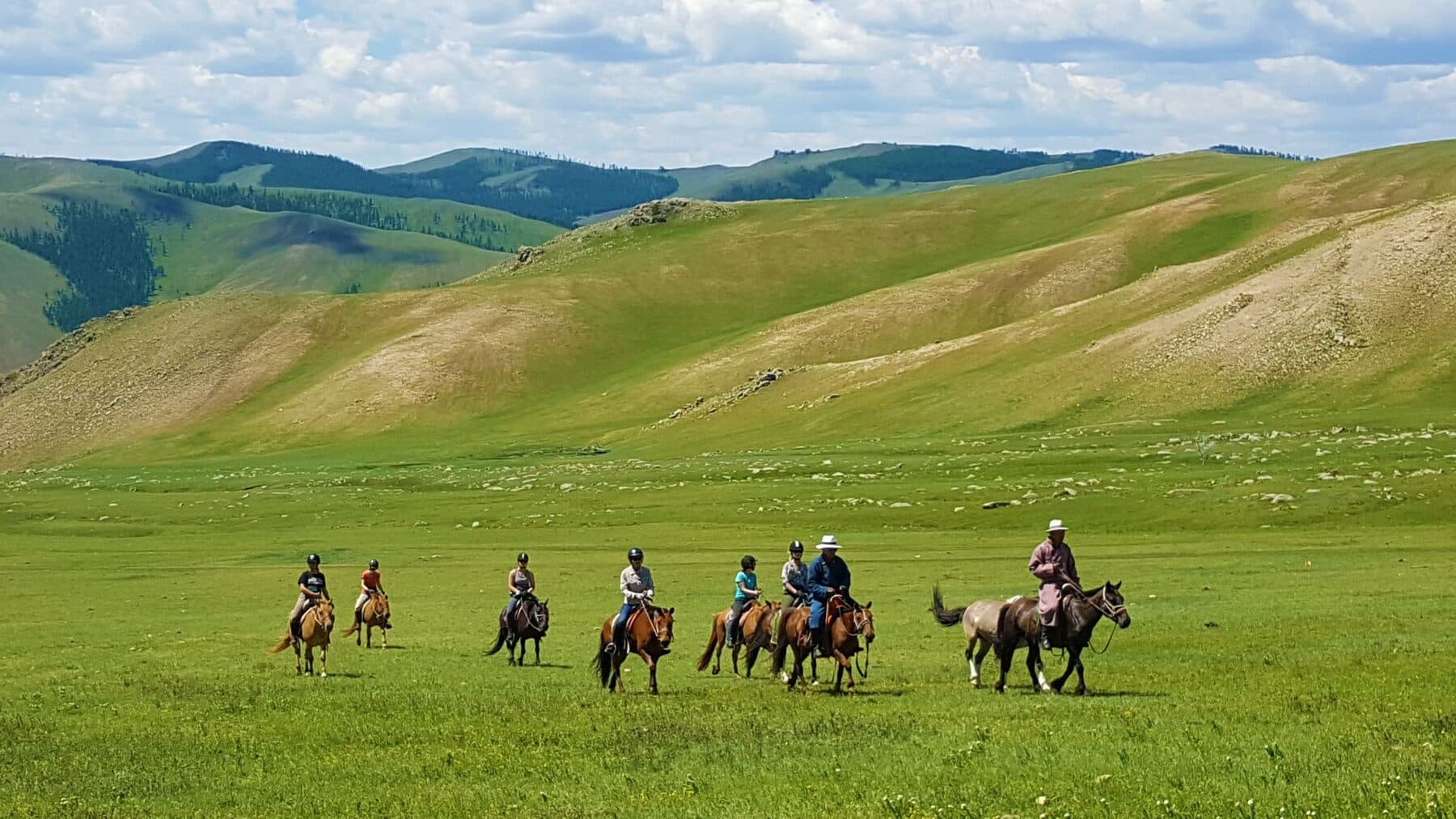 Riders crossing a wide green valley below forested hills