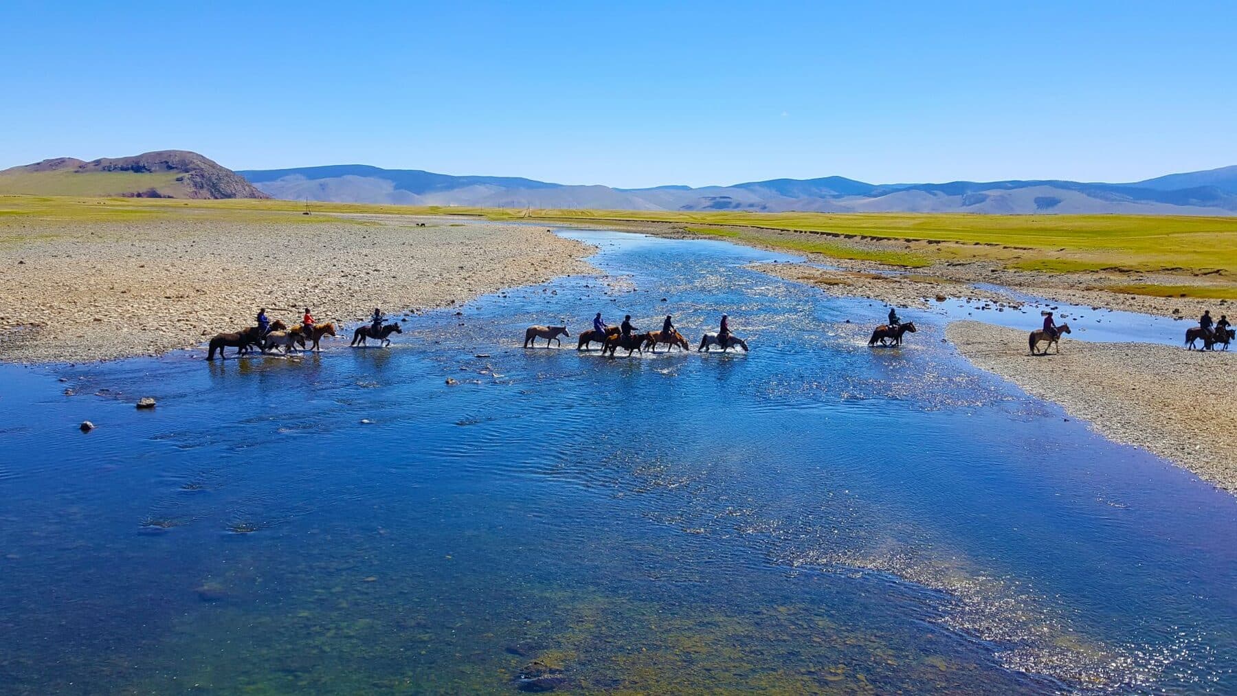 Riders crossing a wide, shallow river in a valley